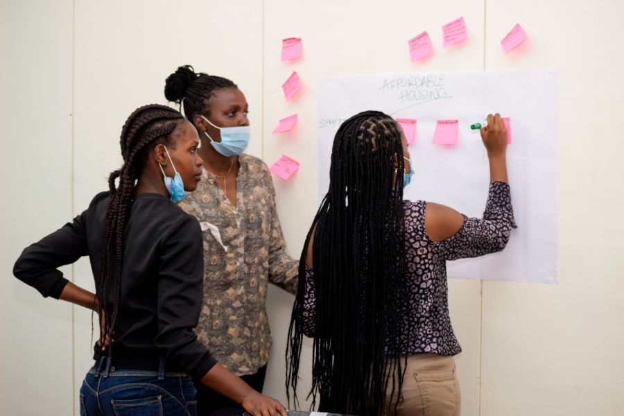 A participant writing on a sticky note mounted on an idea board while her two other team mates look on, during the Better Cities Challenge boot camp